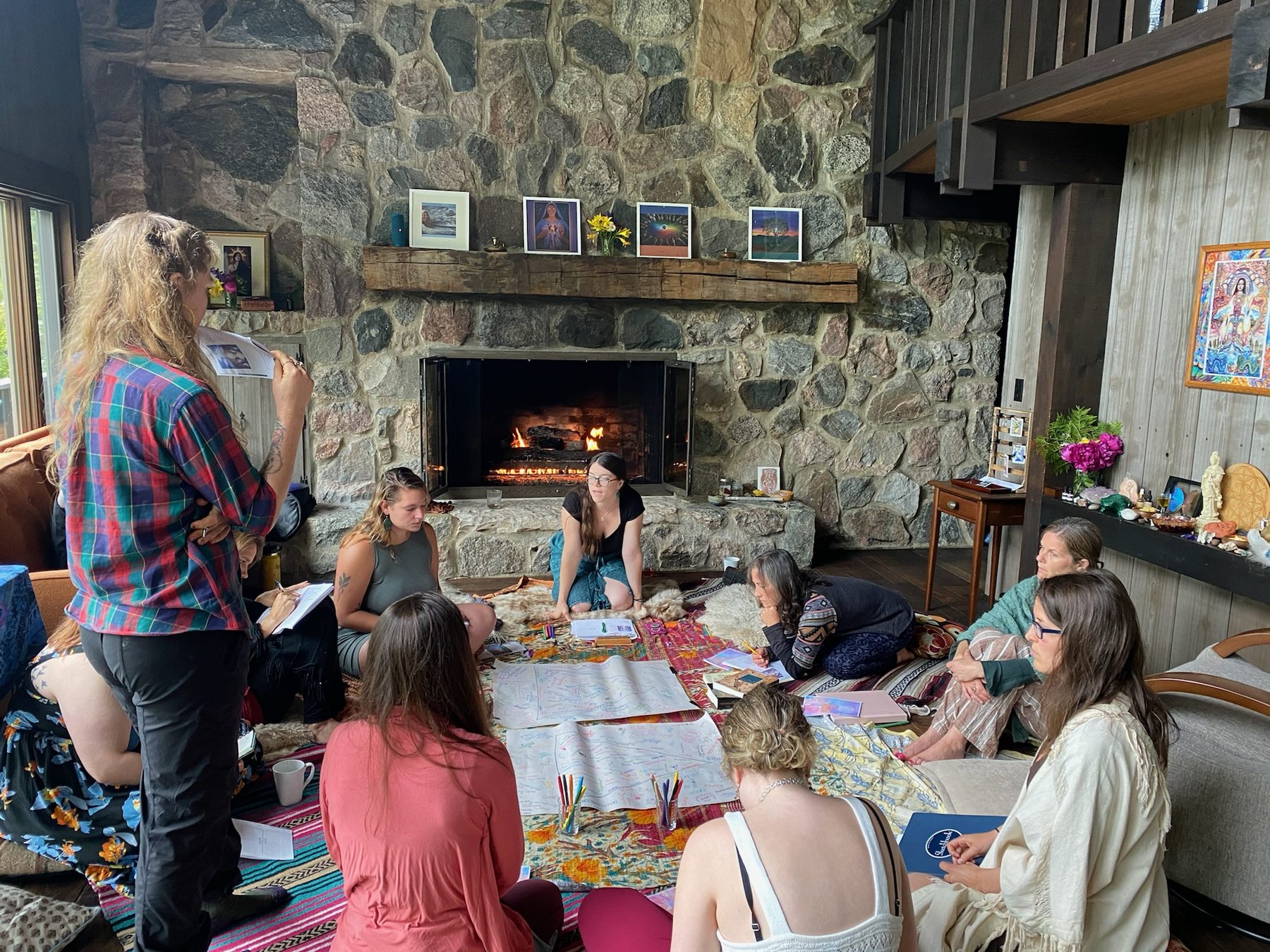 A group seated in a circle on rugs and sheepskins in front of a stone fireplace, working over large sheets of paper while Gabrielle teaches